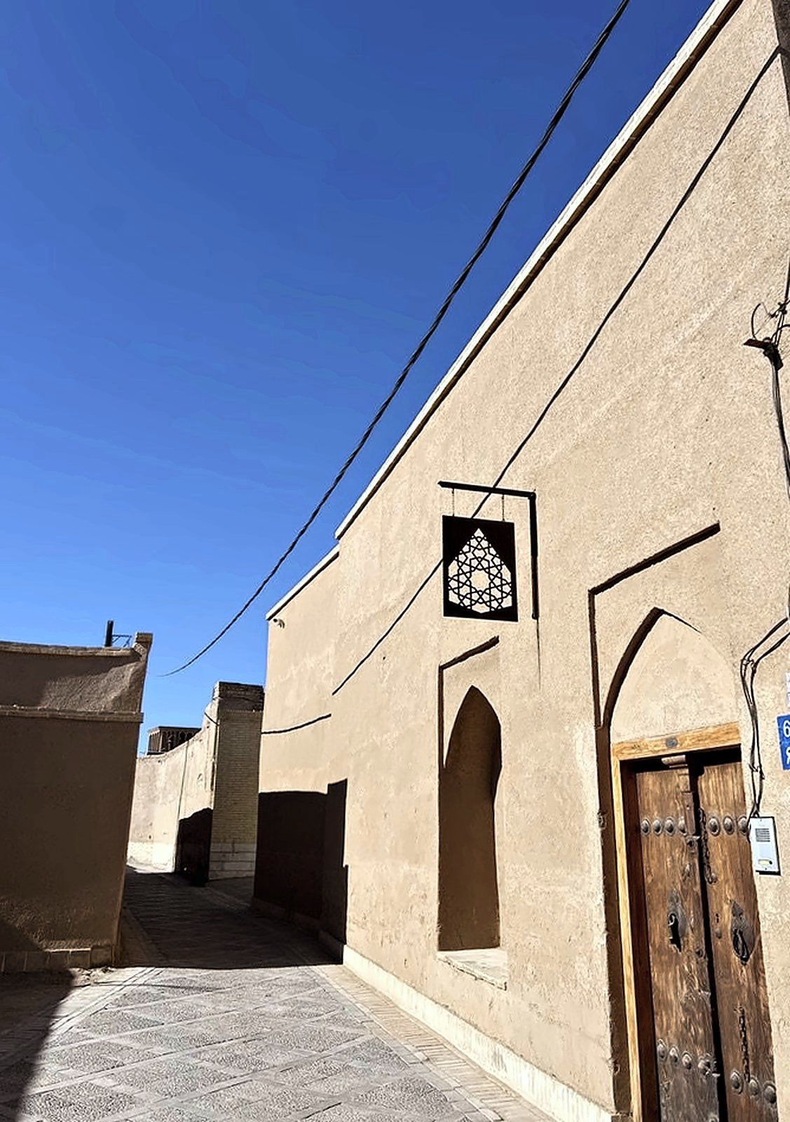 The kucheh outside Lari House: earthen walls, a pointed arch and the house sign under a deep blue sky